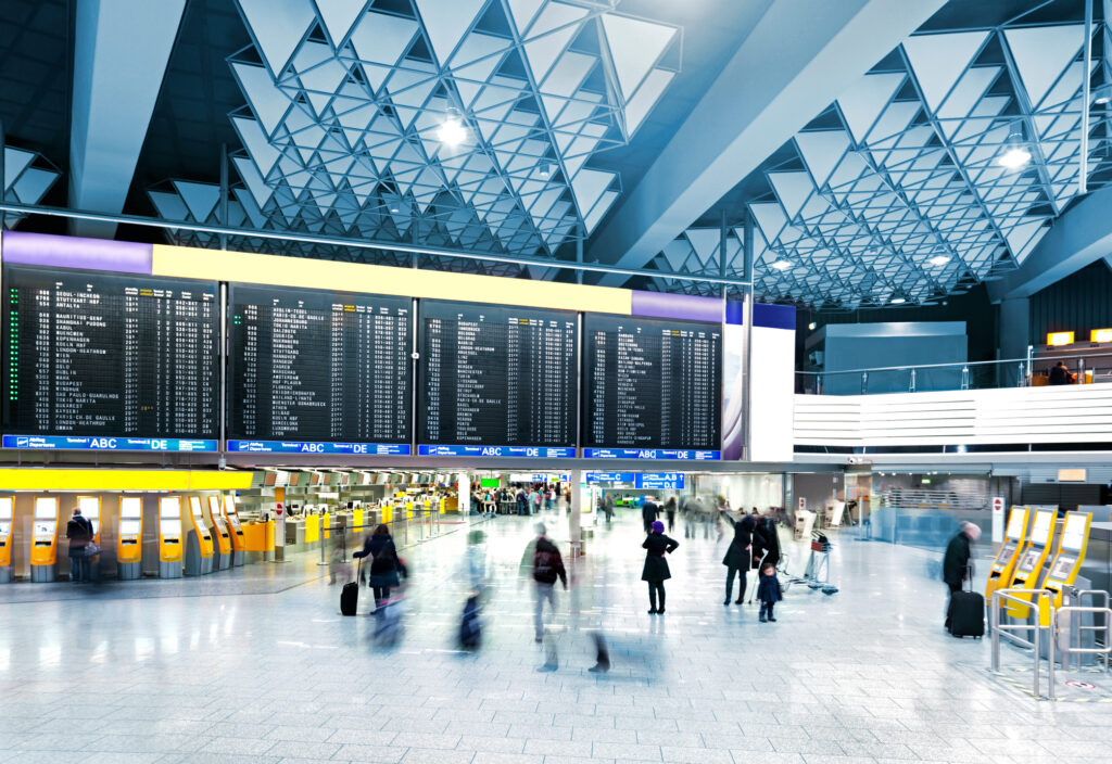 Airport terminal concourse with overhead paging speakers and flight information displays, featuring an adaptive sound system that stays intelligible amid changing background noise.